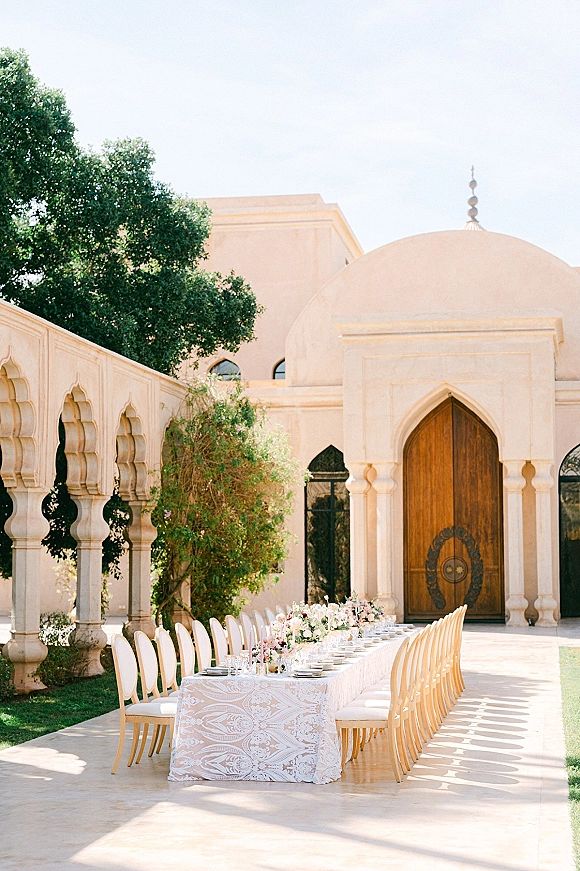 Outdoor reception table set with white linens, floral centerpieces, and glassware, stretching under an arched colonnade courtyard backdrop