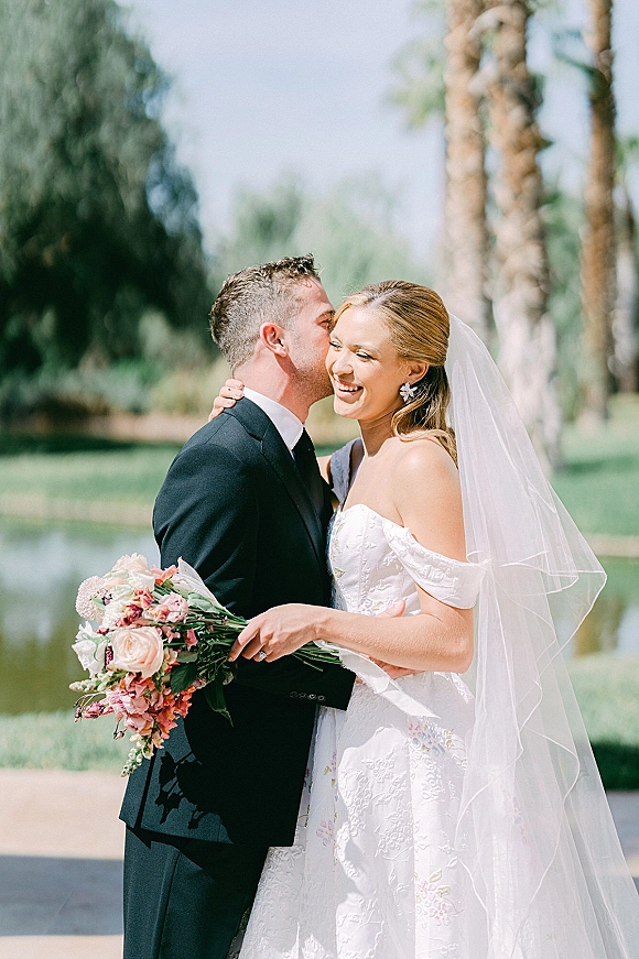 Couple portrait of groom kissing bride’s cheek as she laughs, holding her bouquet on a lakeside lawn with palm trees behind them
