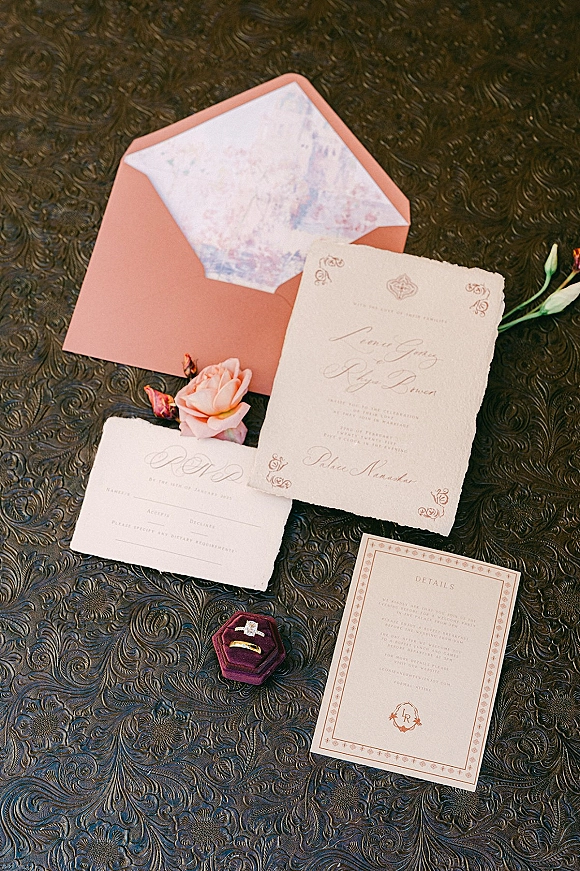 Invitation suite wedding stationery flat lay with deckle edge cards, blush envelope, wax seal, rings and rose on embossed table surface