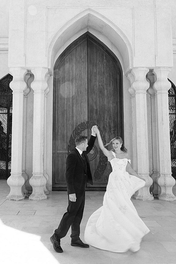 Couple portrait of bride twirling photo with groom leading her spin, dress swirling by an arched doorway and grand wooden doors, earrings gleaming