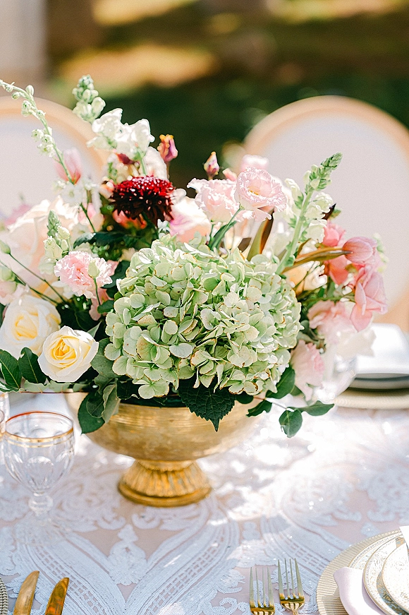Wedding centerpiece with hydrangea centerpiece blooms in a gold compote vase on a lace tablecloth with gold flatware and outdoor greenery behind