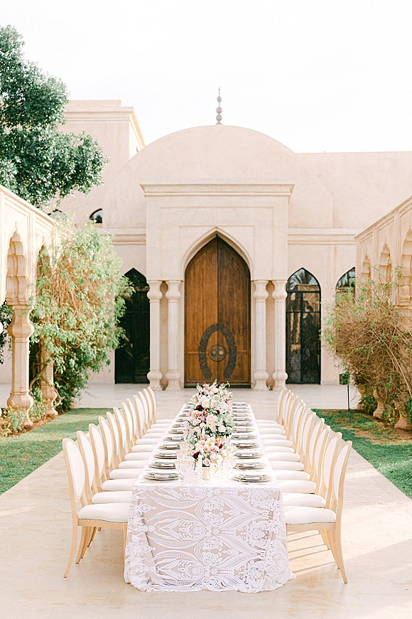 Reception tablescape with long banquet table setup on a white lace tablecloth, floral runner, and place settings in an arched courtyard doorway setting
