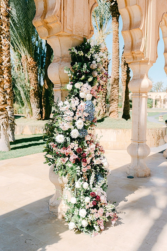 Wedding floral installation with pastel roses and hydrangea garland draped on stone columns beneath arches, with palms and blue sky