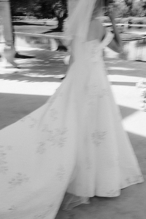 Bride walking away in a strapless wedding dress with a flowing cathedral veil along a garden walkway by trees and a pond