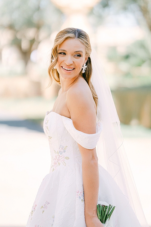 Bridal portrait of a smiling bride in a strapless wedding dress with a long veil, drop earrings, and bouquet against green trees and lawn