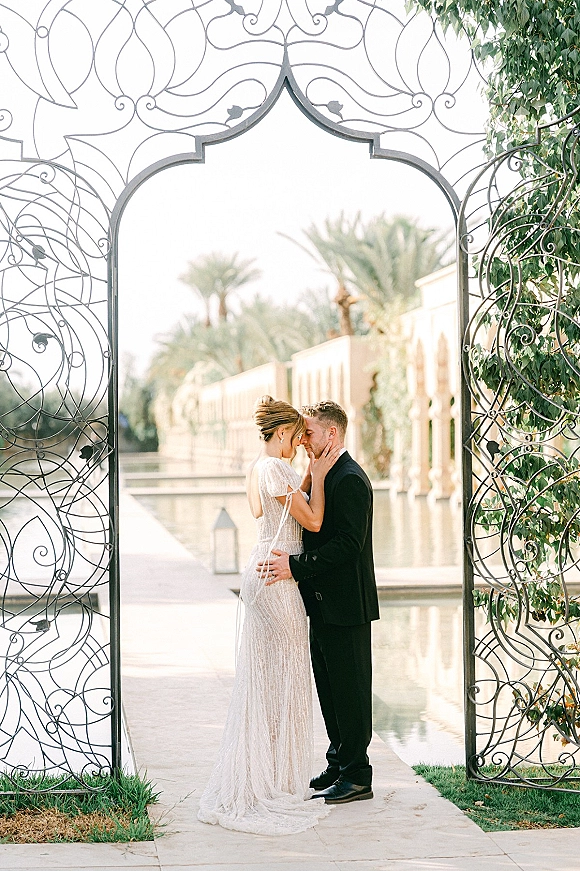 Wedding couple portrait of bride and groom embrace with forehead touch by a reflecting pool, beaded long-sleeve gown at wrought iron gate lanterns