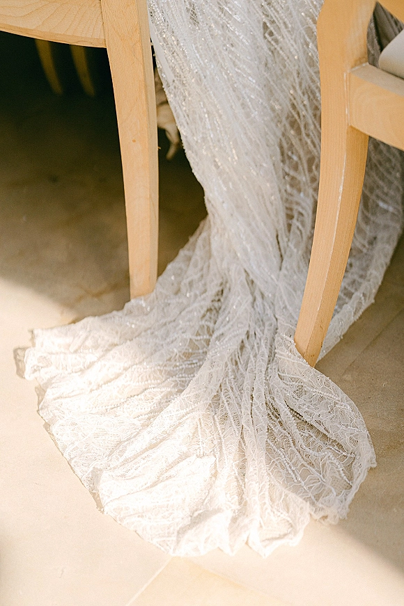 Wedding dress train with lace fabric and beaded embellishments draped over a wooden chair on a tiled floor indoors