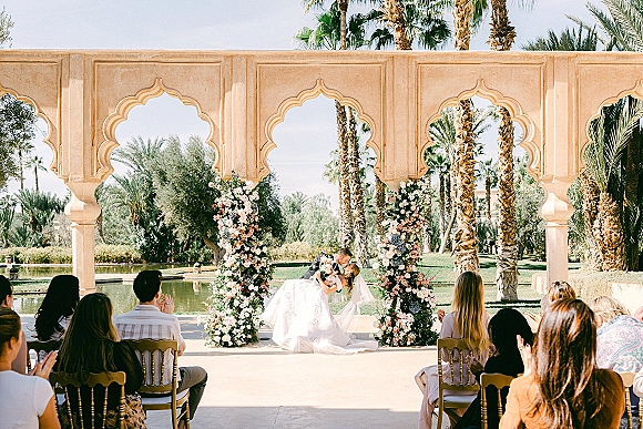 Ceremony kiss moment as the bride and groom dip-kiss beneath floral arch pillars, veil flowing at an arched pavilion by a reflecting pool