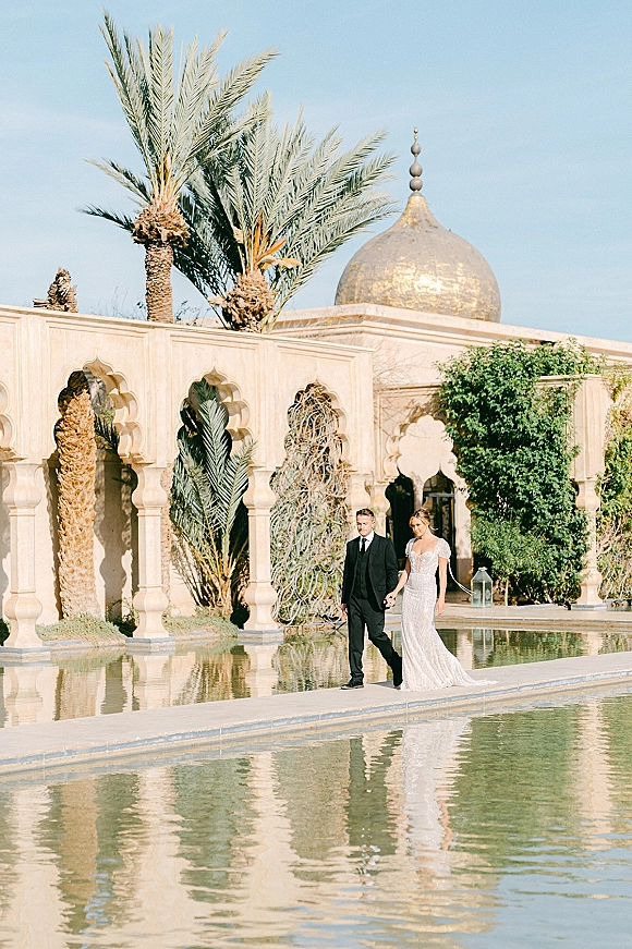 Couple portrait of bride and groom walking hand in hand by a reflecting pool, her veil trailing under stone arches and palms in a courtyard