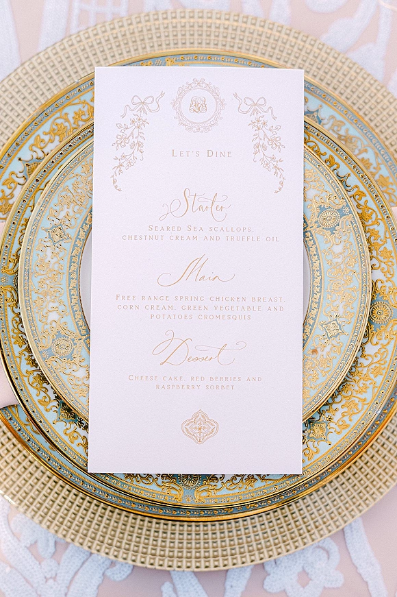 Wedding menu card with gold calligraphy and monogram crest on an ornate blue and gold charger over a blush tablecloth and lace runner