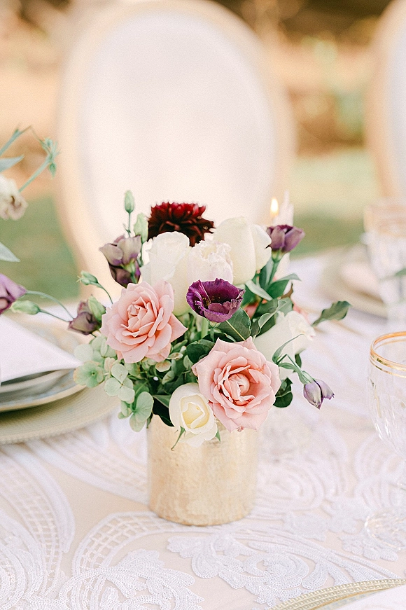 Reception centerpiece of blush and purple roses with greenery in a stone vase on lace tablecloth, candlelit outdoor lawn setting with bokeh chairs