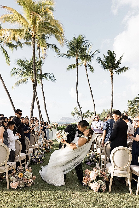 Wedding kiss as groom dips bride in lace gown and veil, bouquet in hand, leis and aisle florals by oceanfront palms and chairs
