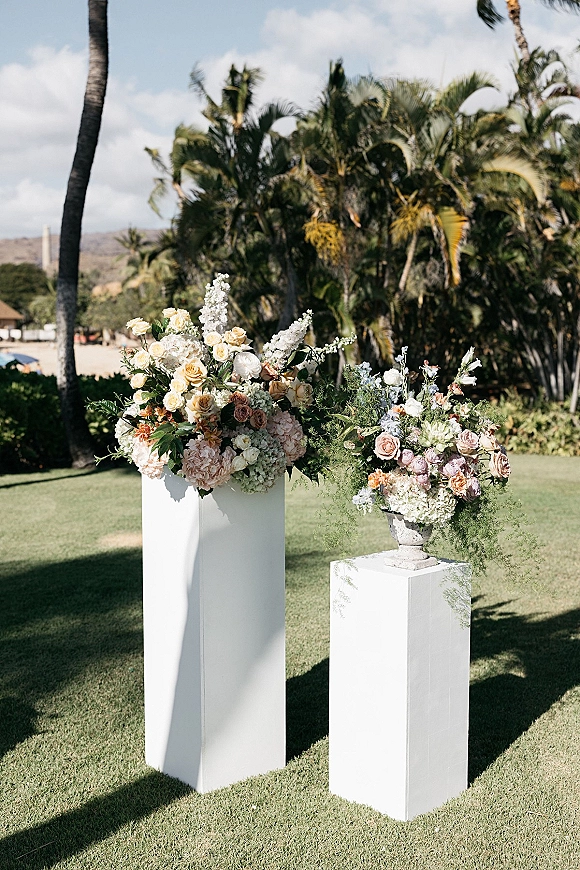 Wedding floral arrangements of pastel roses, hydrangeas, and delphinium with greenery on white plinths and a stone urn on a palm-lined lawn
