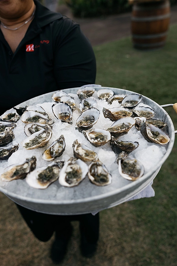 Oyster bar with oysters on the half shell on crushed ice in a metal tray, served by a uniformed caterer on a grass lawn near a barrel