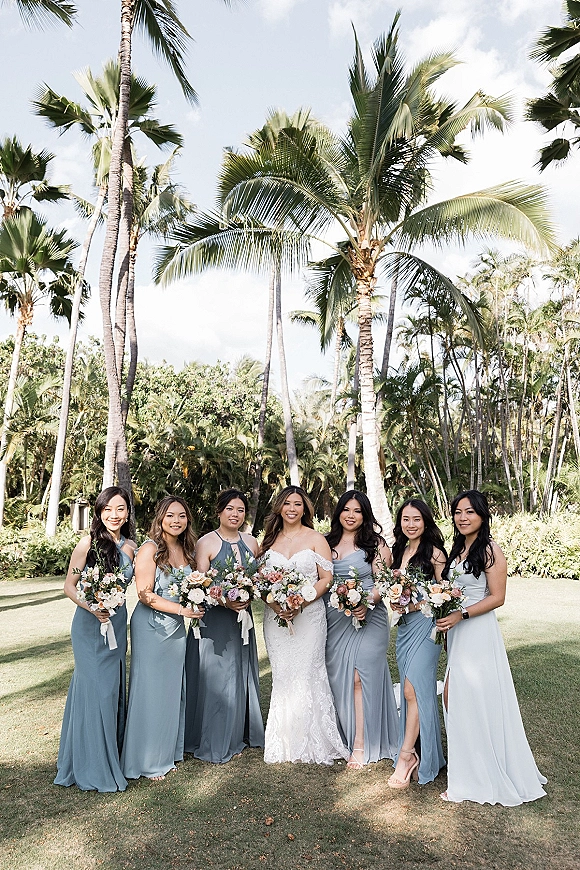 Bride with bridesmaids in a bridesmaids group photo, holding pastel bouquets in dusty blue dresses on a tropical lawn with palm trees