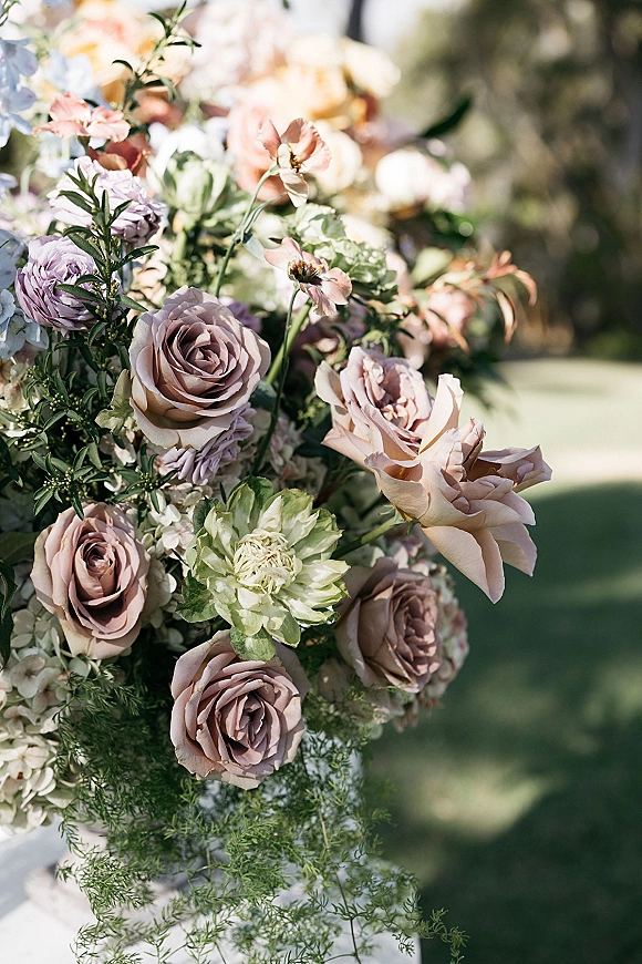 Wedding floral arrangement with roses and mixed blooms, lush greenery, set on a sunlit outdoor lawn with trees in the background