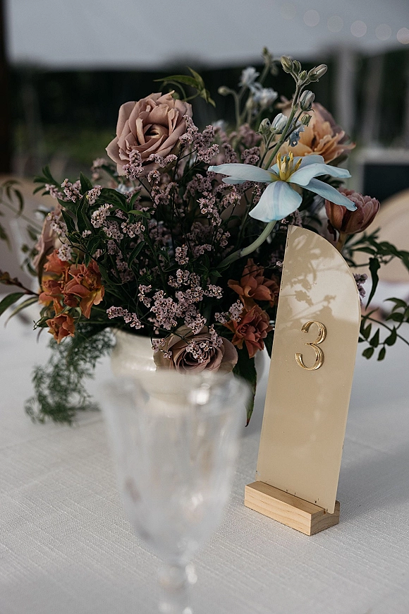 Wedding table centerpiece with wildflower blooms and roses beside a gold table number on linen, set on an outdoor patio with bokeh lights