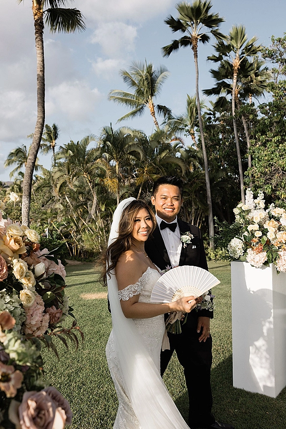 Couple portrait of bride in strapless lace gown and veil holding bouquet and fan beside groom in black tuxedo, palm garden backdrop