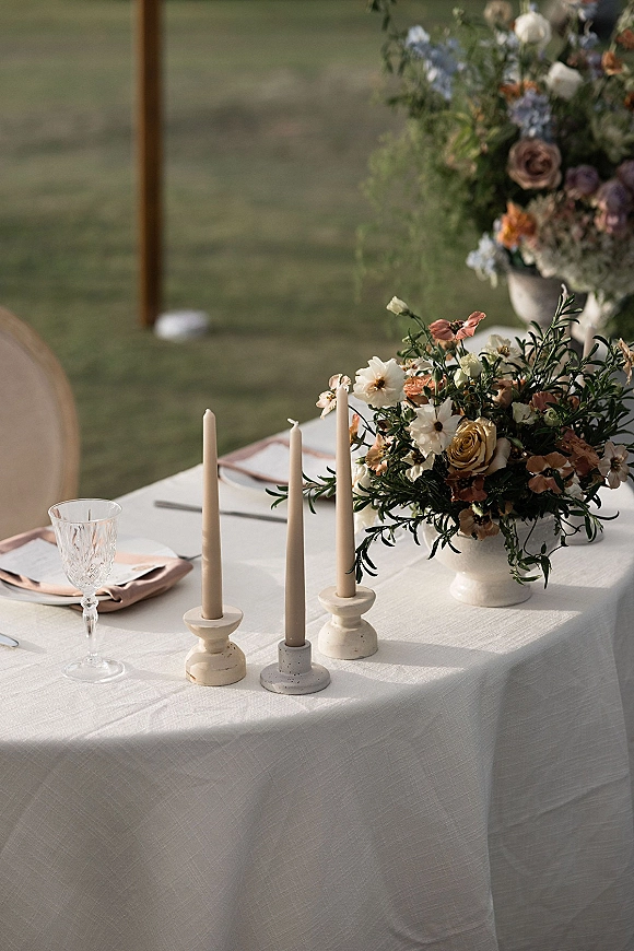 Reception tablescape with wedding table centerpiece in a ceramic vase, neutral taper candles, crystal glassware, and place cards on a lawn outdoors