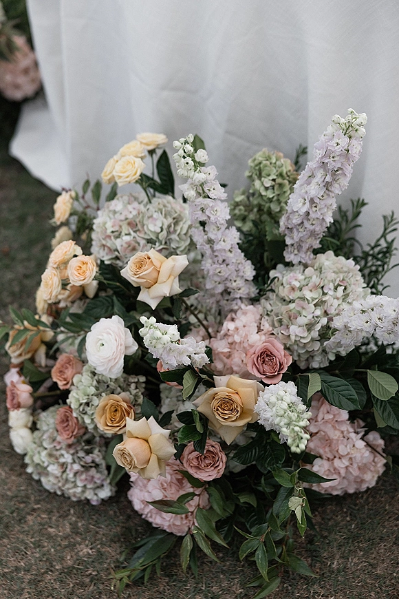 Wedding floral arrangement of roses and hydrangeas with greenery on grass beside a white fabric drape, creating a soft ceremony ground floral cluster
