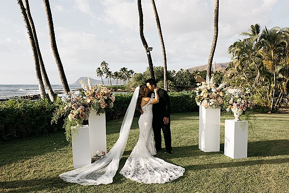 Wedding kiss portrait of bride and groom kissing, her long veil flowing beside floral pedestals on a lawn with palm trees and ocean beyond