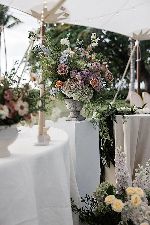 Wedding floral arrangement in a stone urn with pastel roses, hydrangea, and greenery on a white pedestal under a tent with string lights