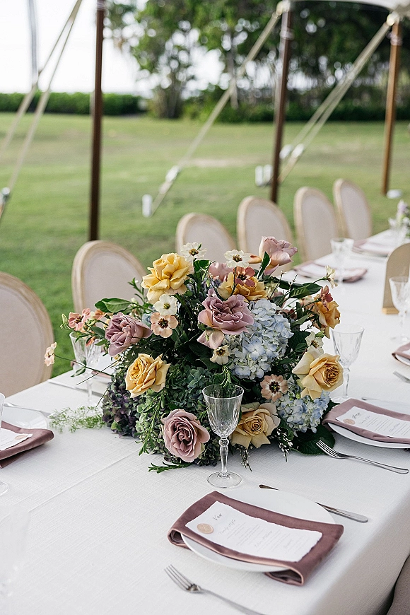 Reception tablescape with roses and hydrangea centerpiece on a long white linen banquet table, mauve napkins and goblets under a lawn tent