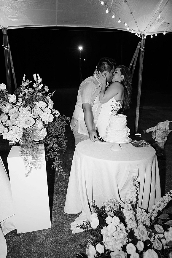 Wedding cake kiss as the newlyweds lean over a two-tier cake on a floral table, bride’s hand on groom’s face under string lights in a tent at night