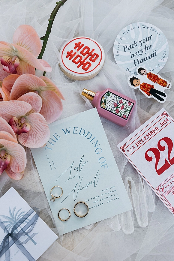 Wedding invitation suite flat lay with blue wedding invitations, gold bands, engagement ring, orchid, perfume, and veil on white tulle fabric