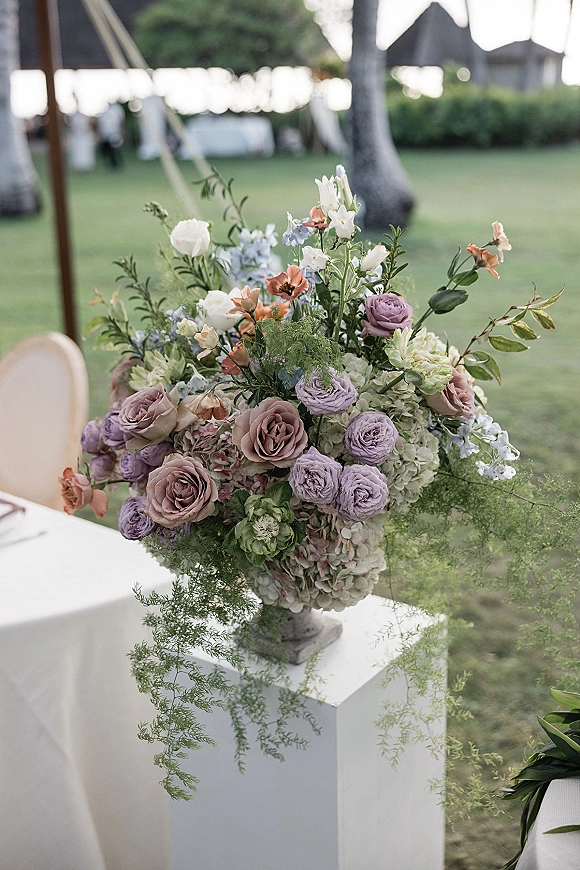 Wedding floral arrangement of roses, ranunculus, and hydrangea with greenery on a white pedestal, set on a lawn near a draped tent and palms