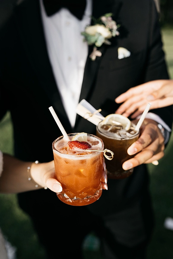 Wedding signature drinks held by a couple in tuxedo, with lemon and strawberry garnishes, paper straws, and ice against blurred greenery