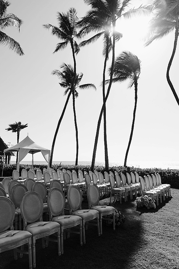 Ceremony setup for a beach wedding ceremony with curved chairs lining the aisle, white canopy and florals on a lawn by ocean and palms