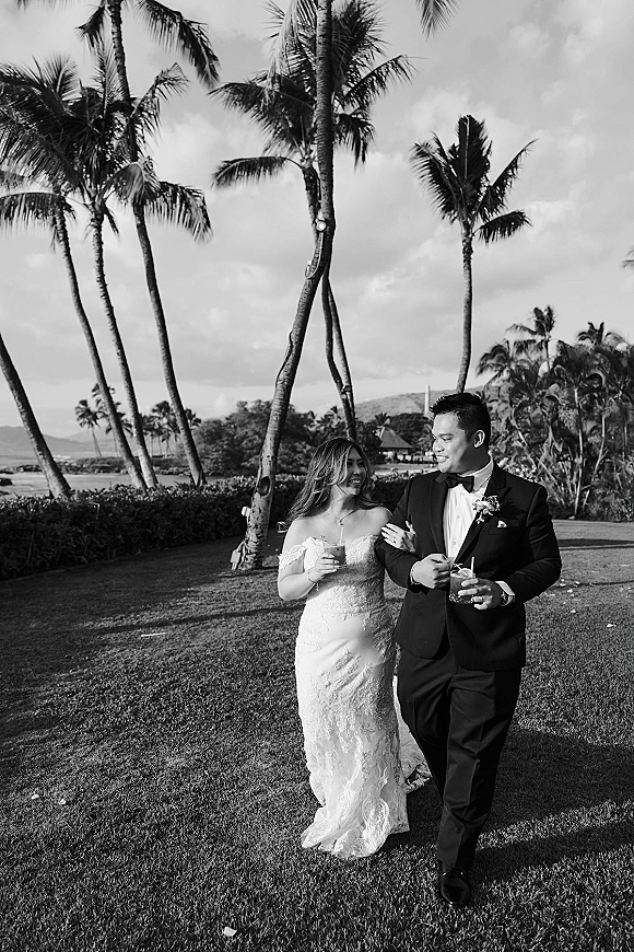 Couple portrait in a black and white wedding photo, bride in lace dress arm in arm with groom in tux, holding cocktails on a palm-lined lawn by the ocean