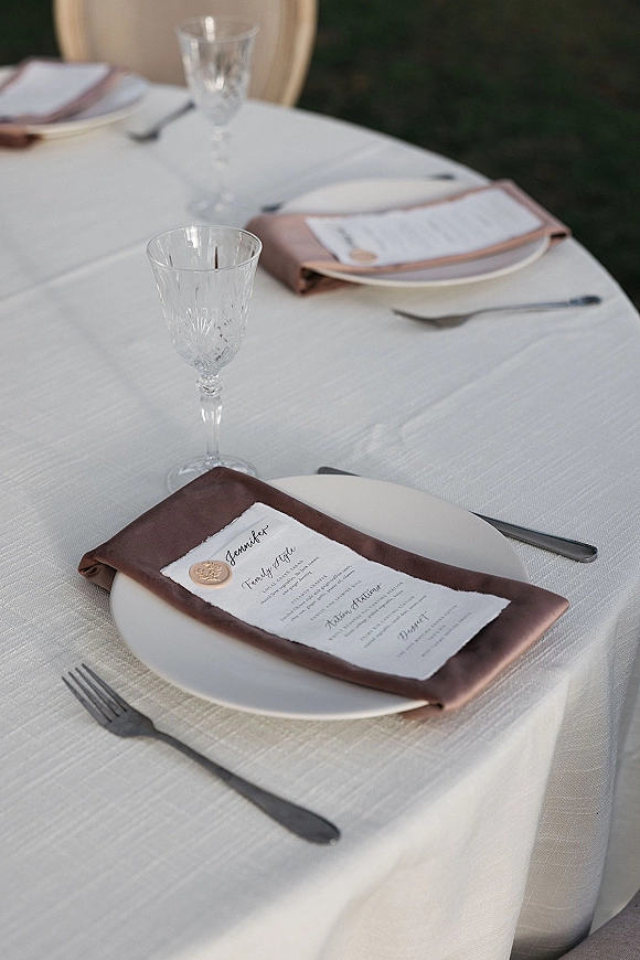 Wedding place setting with brown satin napkins and a wedding tablescape menu card sealed in wax on white plates, crystal goblets, and silver flatware outdoors