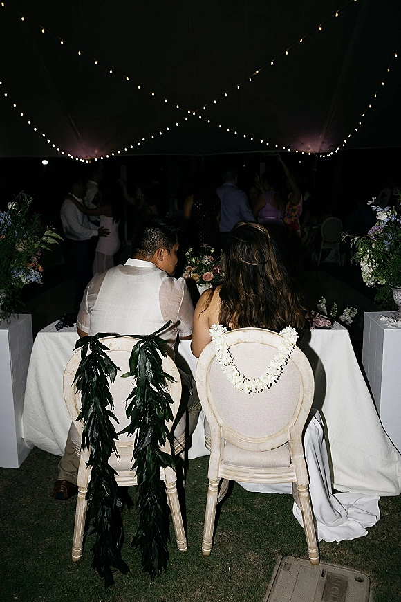 Wedding sweetheart table with string lights canopy, white linen, floral pedestals, and garlanded chairs under an outdoor tent at night
