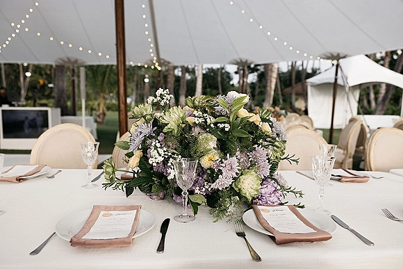 Reception tablescape with wedding table centerpiece of lush florals and greenery, blush napkins, crystal goblets under string lights in a white tent