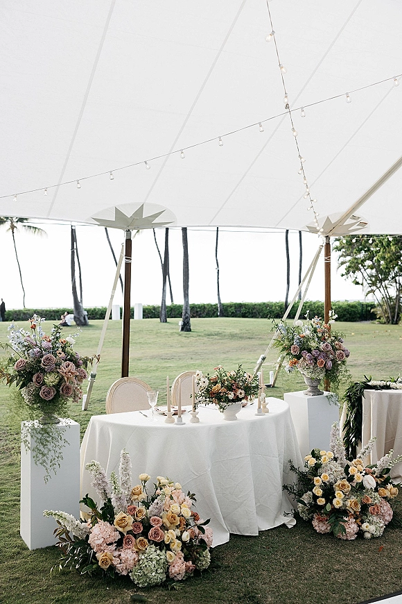 Wedding sweetheart table with rose centerpieces and taper candles, set under a white tent canopy with string lights and palm trees beyond