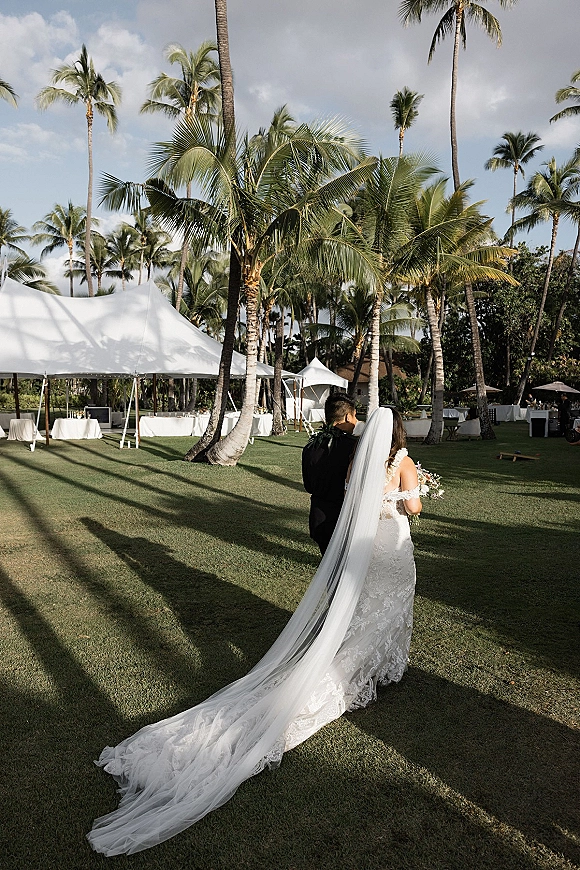 Wedding couple portrait of bride and groom from behind, walking on a tropical lawn with palm trees and a white tent, veil train flowing