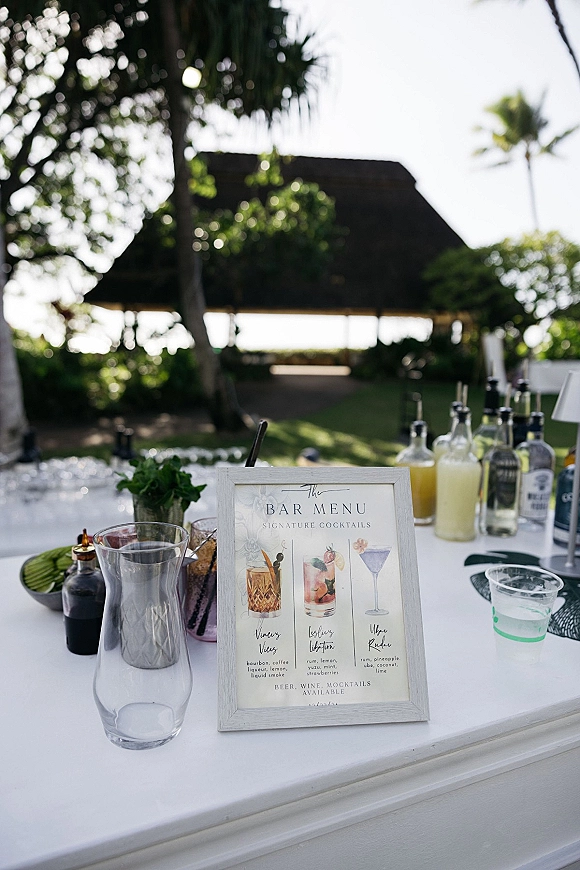 Wedding bar menu on a white cart with a framed sign, liquor bottles and glassware, set on an outdoor lawn with palm trees