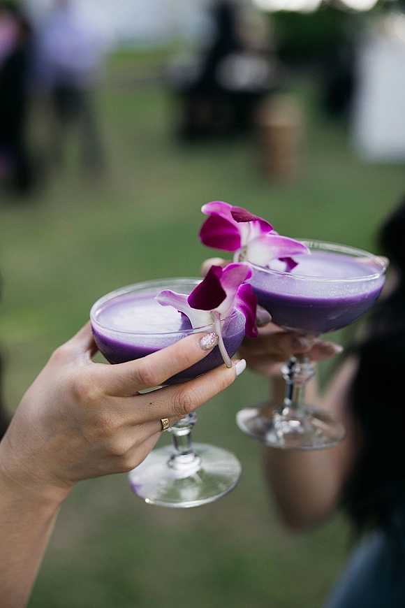 Signature wedding cocktails in coupe glasses, purple drinks with orchid garnish, held by manicured hands with ring on an outdoor lawn