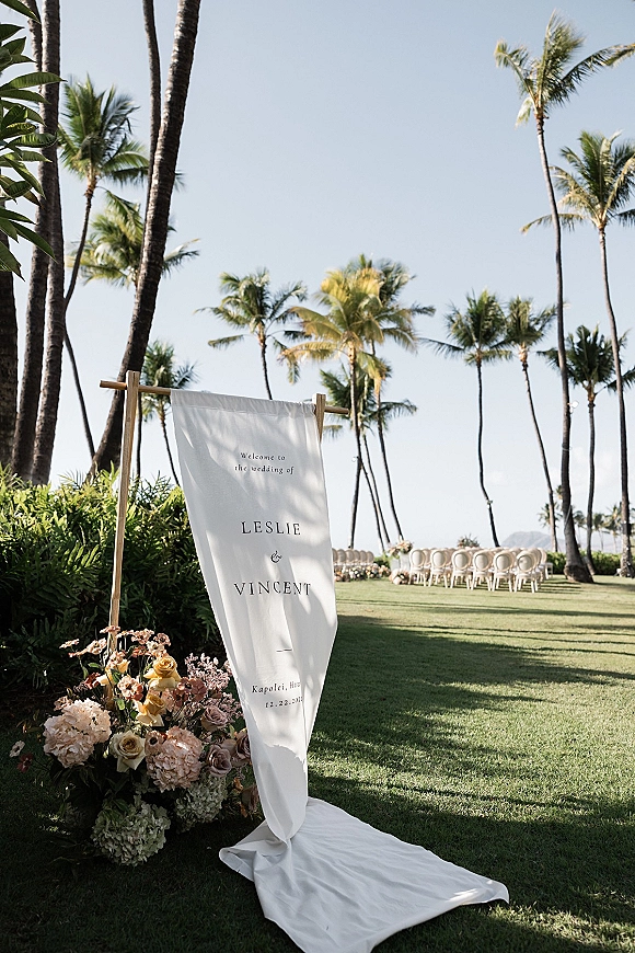 Wedding welcome sign on a wooden stand with a hanging fabric banner and blush rose-hydrangea florals by palm-lined lawn chairs