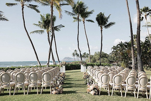 Outdoor ceremony setup for an oceanfront wedding ceremony with floral-lined aisle, white pedestals, and chairs on a palm-lined lawn by the sea