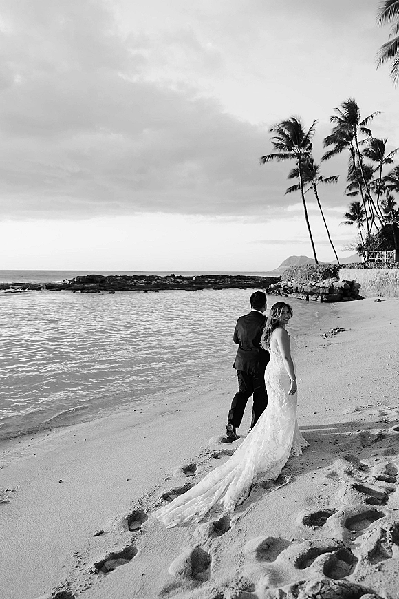 Couple portrait of bride and groom walking on a beach wedding couple shoreline, her lace dress train trailing in sand under a cloudy sky