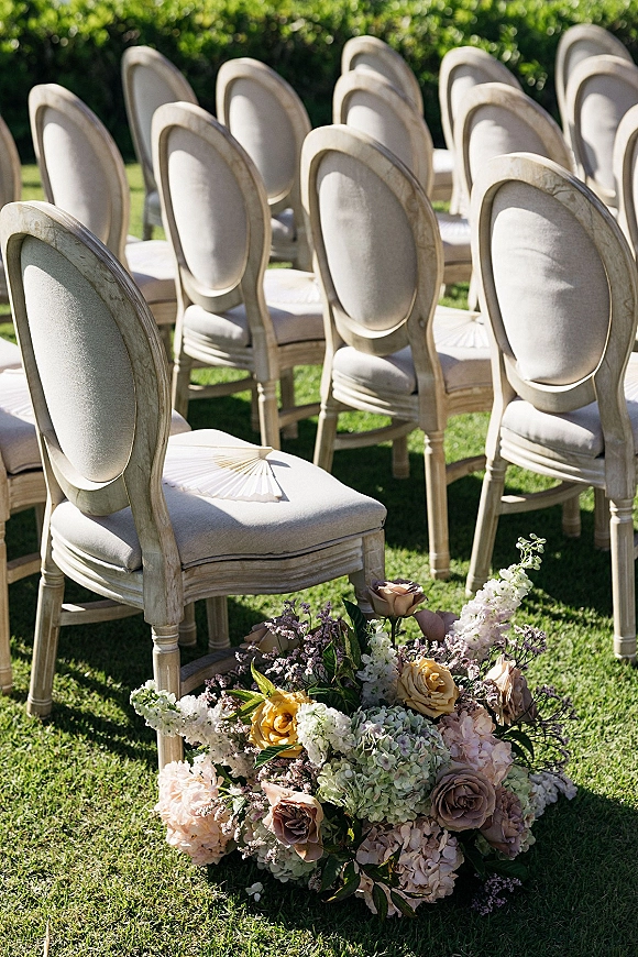 Ceremony seating with outdoor ceremony chairs in rows on a sunlit lawn, oval-back upholstery, hand fans, and low rose-hydrangea florals