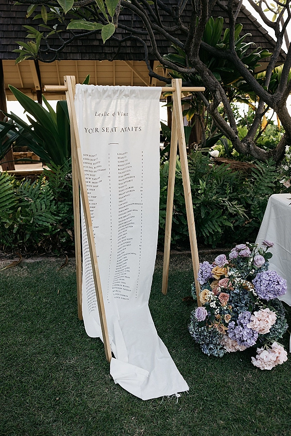 Wedding seating chart on a fabric seating chart banner displayed on a wooden stand with rose and hydrangea florals on a grassy lawn under a pavilion roof