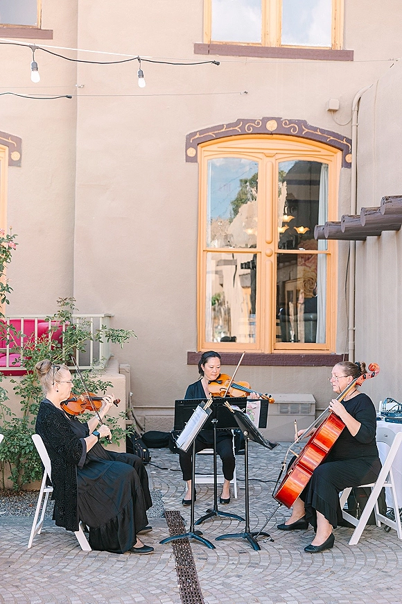 Wedding musicians, a wedding string trio in black playing violins and cello with sheet music on stands in a courtyard patio under string lights