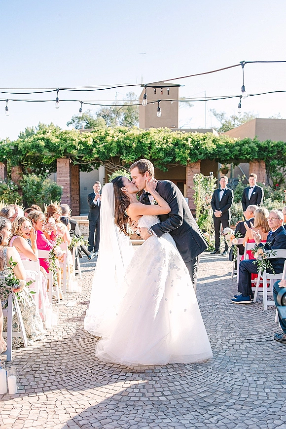 Wedding kiss as the couple shares a ceremony kiss moment on a stone aisle with greenery, string lights overhead, and guests in a brick courtyard