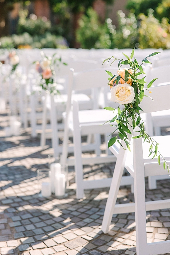 Ceremony aisle decor with white chair ceremony setup, roses and greenery garland, and candle clusters lining a stone paver garden path
