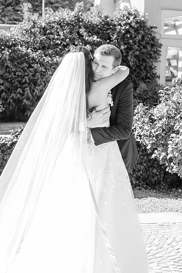 Wedding hug as bride and groom embrace, her veil draped over a strapless dress, his boutonniere visible by hedge greenery and brick walkway