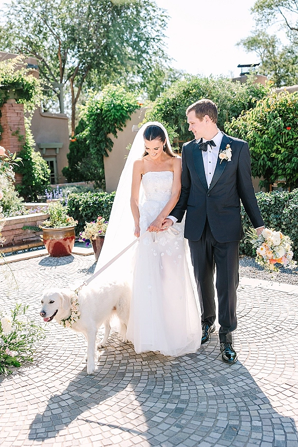 Couple portrait of bride and groom with dog, bride in strapless dress and veil holding leash in a garden courtyard with greenery